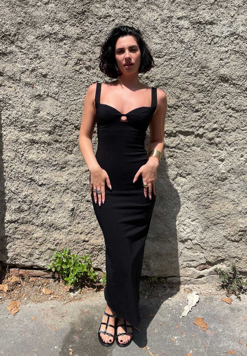 Czarna dopasowana sukienka z dekoltem w kształcie serca i centralnym wycięciem. Gładki materiał, długość do kostek, w połączeniu z płaskimi sandałami na paski i pierścionkami.