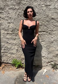 Czarna dopasowana sukienka z dekoltem w kształcie serca i centralnym wycięciem. Gładki materiał, długość do kostek, w połączeniu z płaskimi sandałami na paski i pierścionkami.