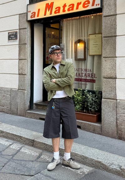 Hombre con gafas, chaqueta verde, gorra negra y pantalones cortos negros largos, de pie con los brazos cruzados fuera de un restaurante llamado "al Matarel."