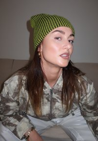 Bonnet en tricot rayé vert et noir porté avec une veste à motif camouflage. Le modèle présente une peau éclatante et de longs cheveux, assis devant un fond neutre.