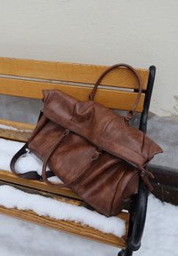 Sac de voyage en cuir marron sur un banc en bois avec de la neige sur l'assise et le sol près d'un mur extérieur.