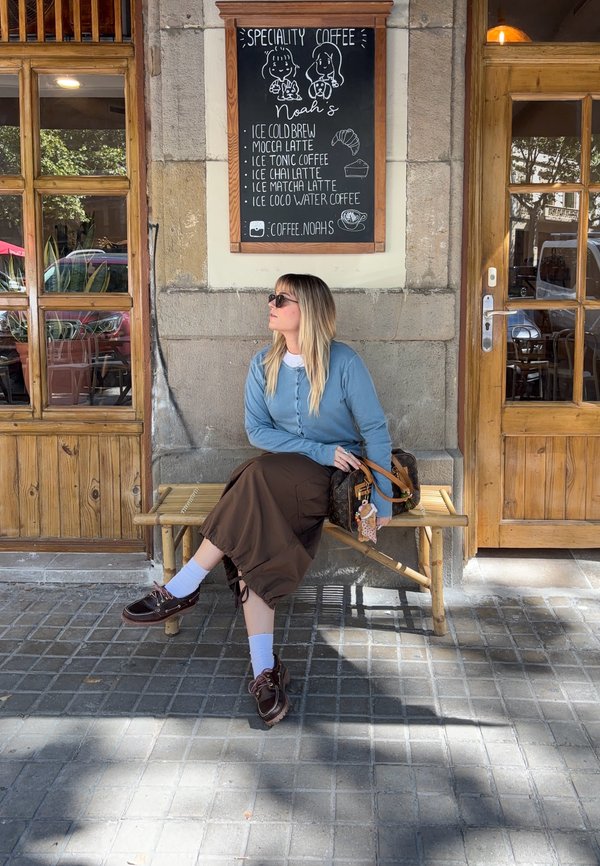 Mujer con gafas de sol sentada en un banco de madera fuera de una cafetería bajo el cartel del menú de café, con una puerta y una ventana de madera en una pared de piedra de fondo.