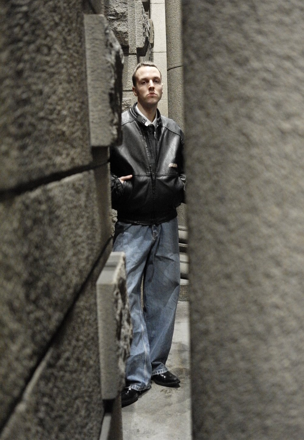 Giacca di pelle nera, camicia bianca, jeans azzurri oversize e scarpe nere; in piedi tra pilastri di pietra grigia con texture.