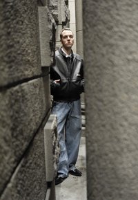 Giacca di pelle nera, camicia bianca, jeans azzurri oversize e scarpe nere; in piedi tra pilastri di pietra grigia con texture.