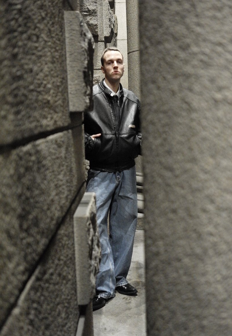 Schwarze Lederjacke, weißes Hemd, übergroße hellblaue Jeans und schwarze Schuhe; steht zwischen strukturierten grauen Steinsäulen.