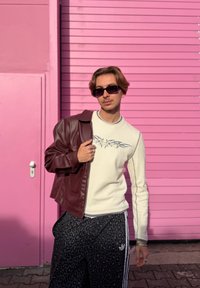 Een persoon draagt een wit, textuur langwerpig shirt met grijs ontwerp, zwarte broek met luipaardprint en een bordeauxrode leren jas tegen een roze achtergrond.