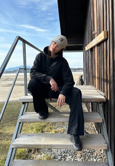 Sudadera negra con detalles grises, pantalones negros y zapatillas oscuras. Gorra de color claro con un logo. Sentado en escaleras de madera.