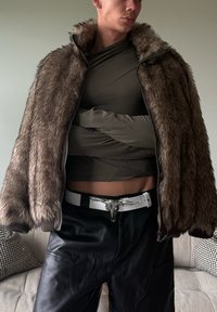 Chaqueta de borreguito marrón con cremallera y cuello alto, combinada con una camiseta ajustada de manga larga color oliva y pantalones de cuero negros, que cuentan con un cinturón blanco.