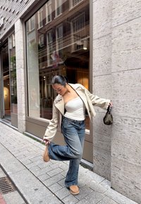 Chaqueta corta de un beige claro, camiseta blanca sin mangas, jeans acampanados azules y zapatillas marrones. Sosteniendo un pequeño bolso estampado mientras está de pie sobre adoquines.