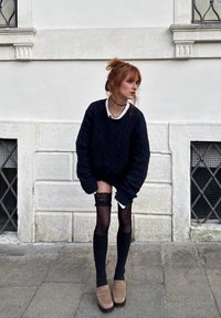 Jeune femme aux cheveux roux coiffés en chignon, portant un pull noir surdimensionné, des bas noirs montants à dentelle et des chaussures à plateforme beige, posant en plein air.