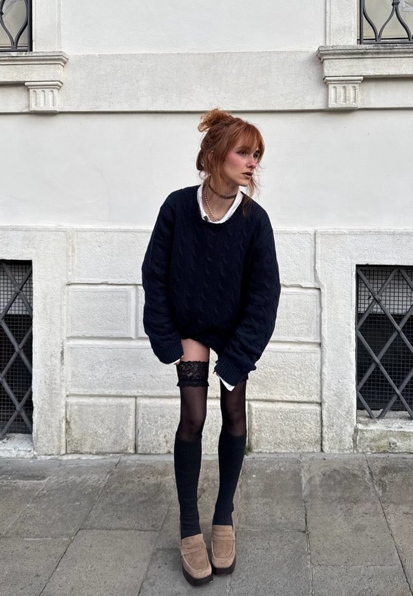 Jeune femme aux cheveux roux attachés en chignon, portant un pull noir oversize, des bas noirs montants avec des hauts en dentelle, et des chaussures à plateforme beiges, posant en extérieur.