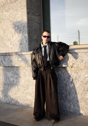 Black leather cropped jacket over a blue shirt and black tie; wide black trousers with pleats; person wearing sunglasses against a stone wall.