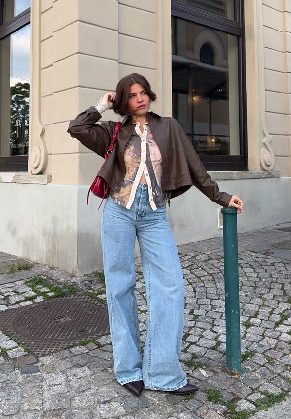 Jeune femme en veste courte marron, chemise à motifs, jeans larges bleus et chaussures pointues, debout sur un trottoir pavé tenant un poteau vert.