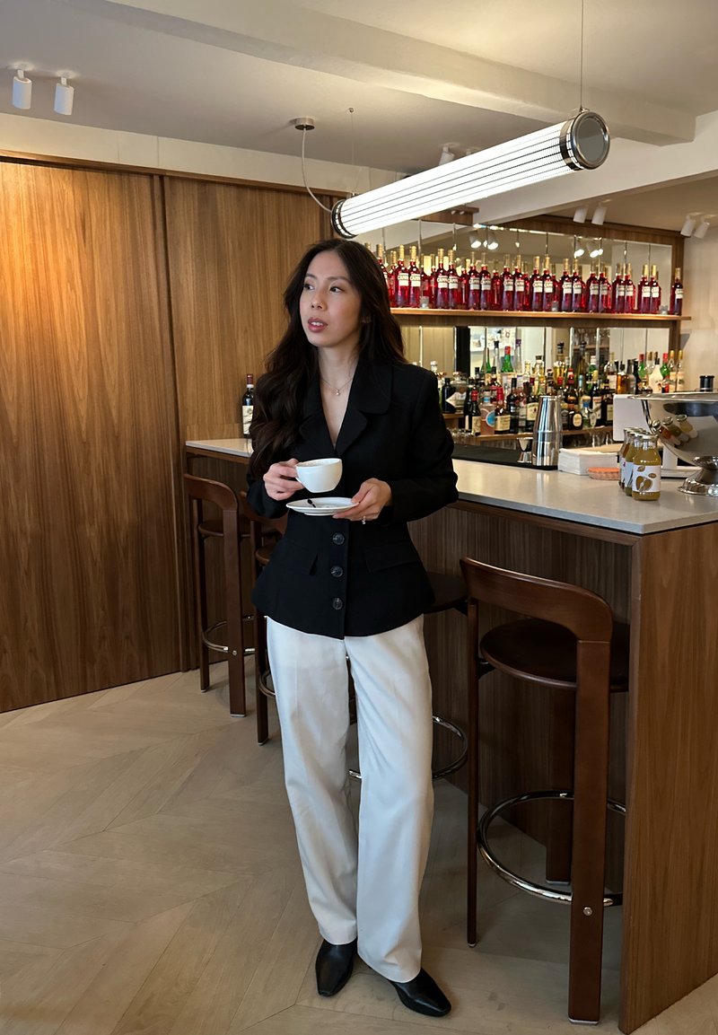 Vrouw in een zwart colbert en witte broek, die een kopje en schotel vasthoudt, staand bij een houten bar met flessen en krukken in een modern interieur.