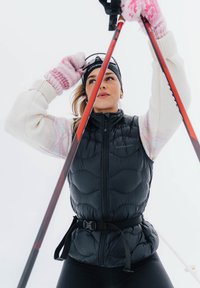 Νεαρή γυναίκα ρυθμίζει τη μαύρη κορδέλα κεφαλής ενώ κρατάει κόκκινους σκιέρ διαστρεμματικού σκι, ντυμένη με λευκό πουλόβερ, μαύρο γιλέκο και ροζ γάντια, με χιονισμένο φόντο.