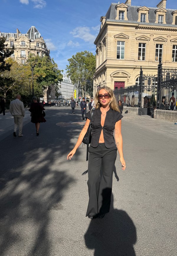 Une femme vêtue de noir marche dans une rue ensoleillée de Paris, avec derrière elle des bâtiments historiques ornés, des grilles en fer, des arbres et des piétons.