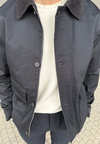 Person som bär en svart jacka med knapp- och dragkedjeförslutningar över en vit skjorta, stående på en kaklad yta.