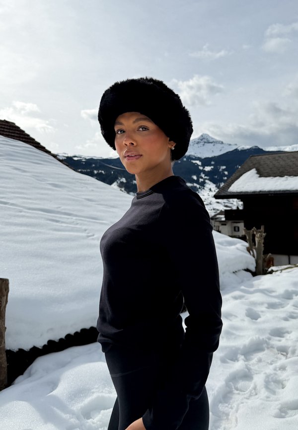 Joven mujer con un sombrero de piel negro y un conjunto negro está de pie afuera en un pueblo montañoso nevado con cielo despejado y nubes dispersas.