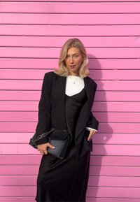 Blazer nero sopra un lungo abito nero con top bianco. Tiene una borsa nera con tracolla a catena. Sfondo a strisce rosa.