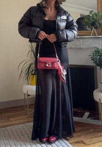 Donna che indossa un piumino nero e un vestito maxi trasparente nero, tiene in mano una borsa rossa, in piedi all'interno vicino a piante e a uno specchio su un camino.