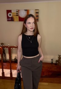 Débardeur noir raccourci, pantalon marron ample, colliers en or superposés et petites boucles d'oreilles anneaux. Tenant un sac noir, debout à l'intérieur.