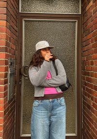 Cardigan gris tricoté sur un haut rose et une chemise marron, associé à un jean bleu à jambes larges. Une casquette grise ornée de décorations et un sac noir.