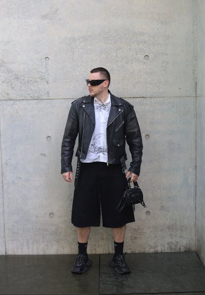 Hombre con chaqueta de cuero negra, camisa blanca estampada, pantalones cortos negros, zapatillas deportivas, gafas de sol y sosteniendo un pequeño bolso negro, de pie contra una pared de hormigón.