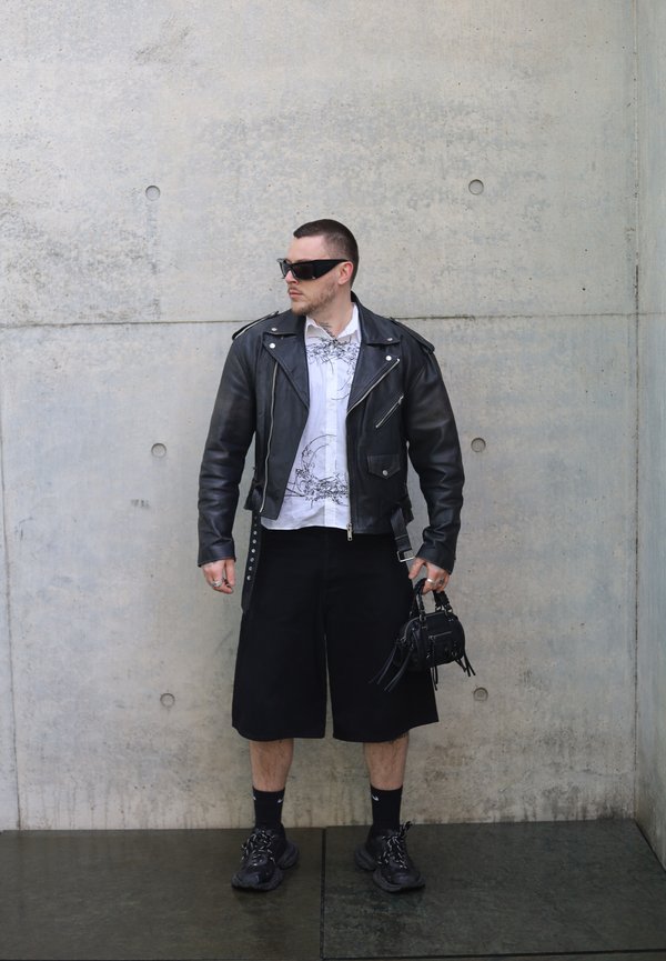 Uomo con giacca di pelle nera, maglietta bianca stampata, pantaloncini neri, sneakers, occhiali da sole e una piccola borsa nera in mano, in piedi contro un muro di cemento.