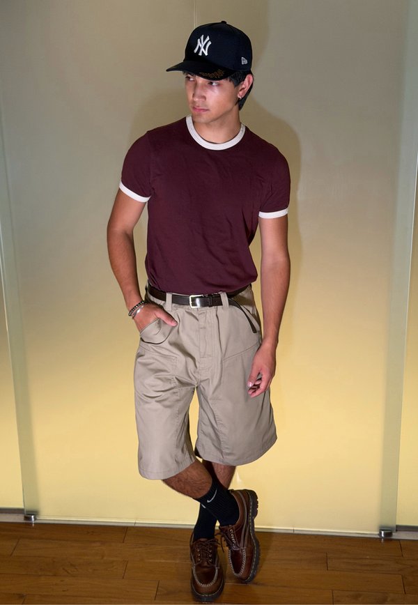 Jeune homme portant une casquette de baseball noire, un t-shirt bordeaux, un short beige, des chaussettes noires et des chaussures marron, debout contre un mur beige.