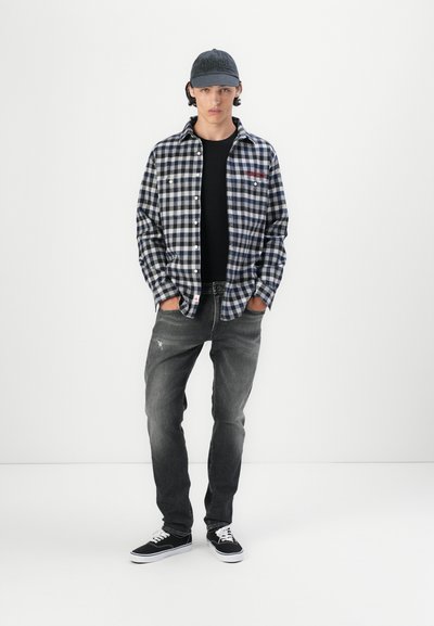 Camisa a cuadros en blanco y negro, camiseta negra, jeans grises y zapatillas negras. Gorra con logotipo. De pie frente a un fondo blanco uniforme.