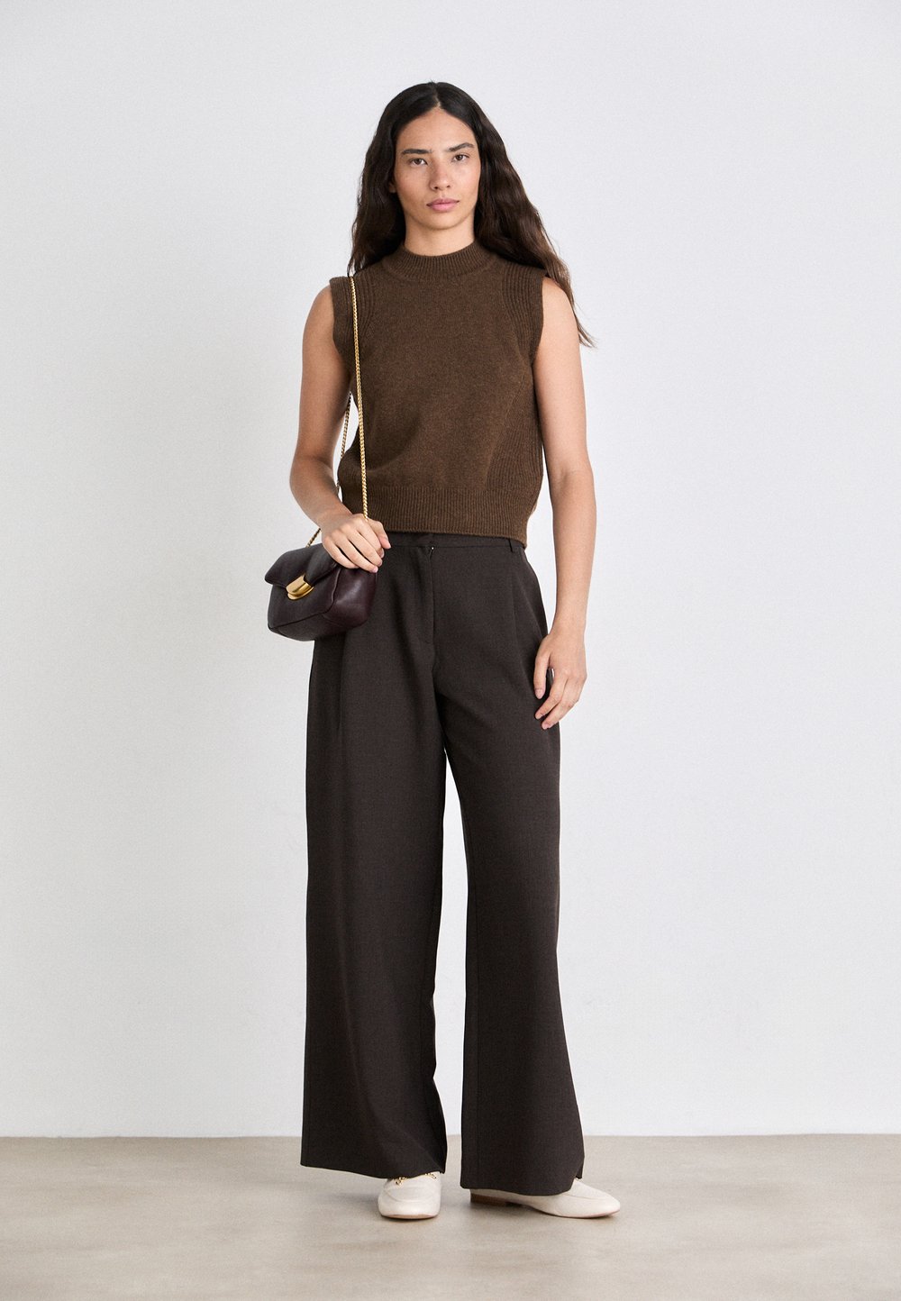 Brown sleeveless knit top teamed with high-waisted, wide-leg dark trousers. Accessories consist of a small burgundy handbag and cream loafers.