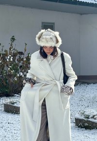 Hvid uldfrakke med bælte i taljen, leopardmønstrede manchetter og en pelsagtig hat; sneklædt baggrund med grønne buske.