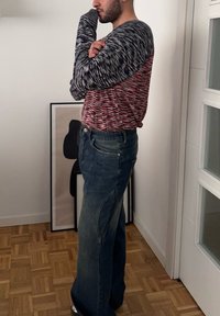 Hosszú ujjú, színes kötött felső piros, fehér és tengerészkék mintákkal; kék, bő szabású farmernadrággal párosítva; egy műalkotás mellett áll.