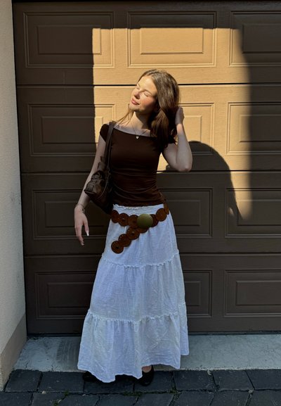 Top marrón ajustado, falda maxi blanca de capas con dobladillo de volantes y un cinturón circular decorativo marrón. El fondo presenta una puerta de garaje marrón texturizada.