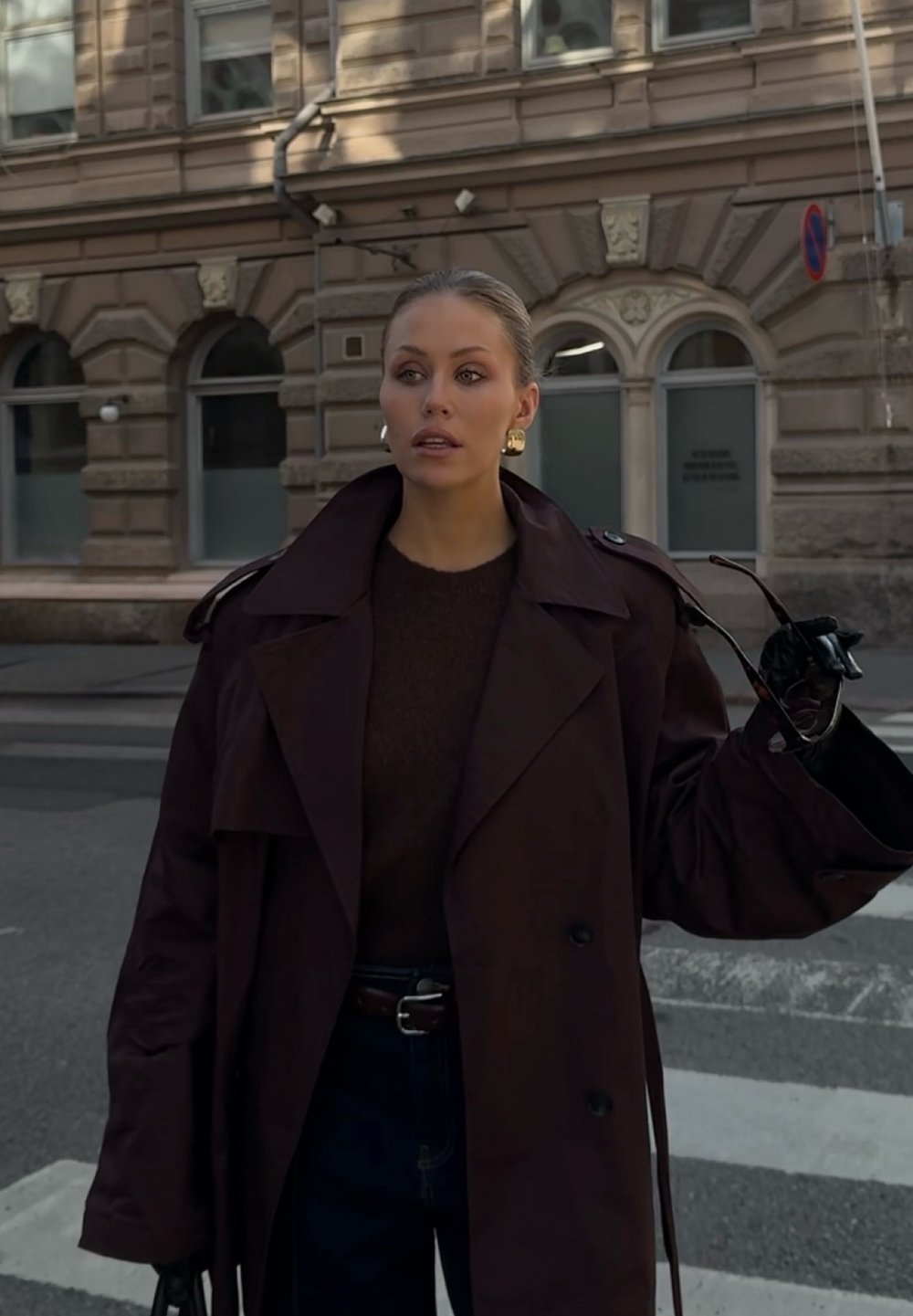 Overdimensjonert mørkebrun trenchcoat med brede revers og dobbelte knapper, sammen med en brun genser og mørke jeans. Holder solbriller.