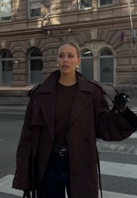 Overdimensjonert mørkebrun trenchcoat med brede revers og dobbelte knapper, sammen med en brun genser og mørke jeans. Holder solbriller.