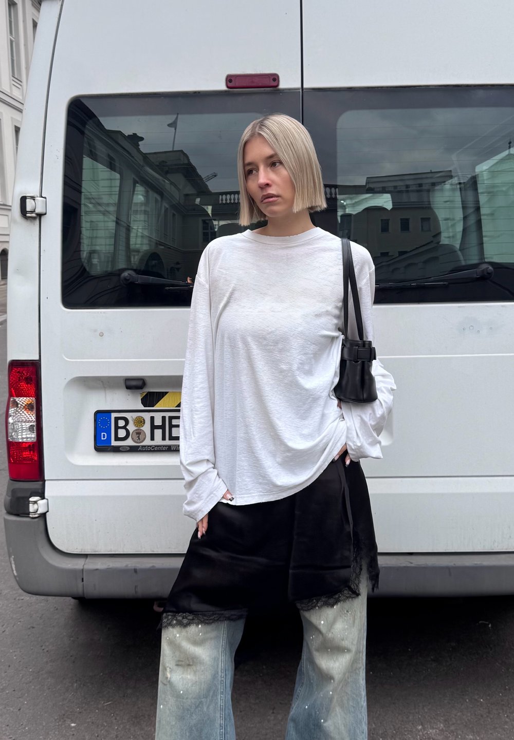 Mujer de cabello rubio corto, vestida con una camisa blanca de manga larga, falda negra y jeans azules, de pie frente a una furgoneta blanca en una calle.