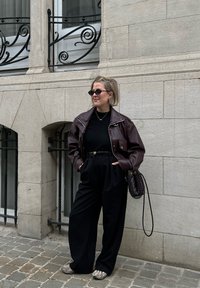 Veste en cuir marron, haut noir, pantalon noir taille haute et chaussures grises. Sac à main noir avec bandoulière tressée. Fond en pavés.