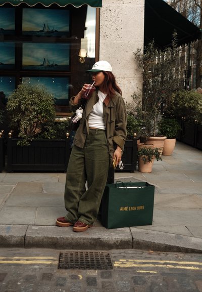 Abrigo oversize verde caqui, top corto blanco, pantalones oliva, zapatos de plataforma marrones, gorra blanca, sosteniendo una bebida, bolsa de compras al lado.