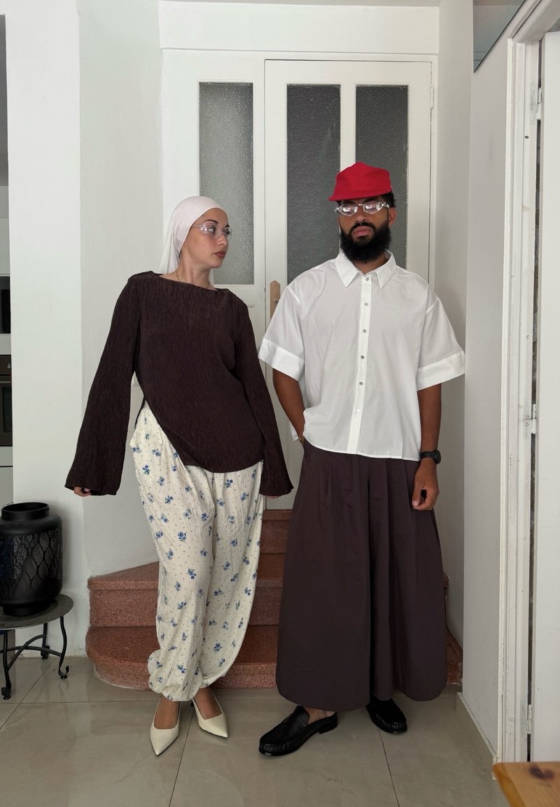 Deux adultes se tiennent à l'intérieur près d'une porte blanche fermée ; l'un porte un foulard blanc et un pantalon à fleurs, l'autre une casquette rouge et un pantalon large et foncé.