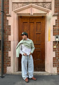 Pulôver verde sobre uma camisa riscada, calças largas de jeans azul claro e sapatos castanhos. Porta de madeira com carvões intrincados e parede de tijolos.
