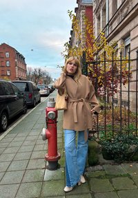 Manteau beige à manches larges et à taille nouée, associé à un jean évasé bleu clair. Hydrant rouge et bâtiments en brique en arrière-plan.