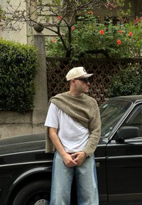 Beige strikkegenser over en hvit t-skjorte, lyseblå vid bukse og en beige caps, står ved siden av en svart bil.