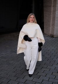 Femme marchant sur un pavé en pierre portant un pantalon blanc, une cape blanche, des chaussures pointues noires, des gants noirs avec une bordure en fourrure, et tenant un petit sac à main blanc.