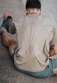 T-shirt de sport beige avec des lignes diagonales au dos, manches courtes, associé à un short vert, sur un fond en béton.