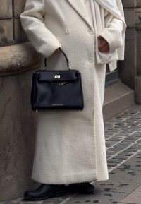 Personne portant un long manteau crème et des bottes noires, tenant un sac à main noir et une écharpe crème assortie, debout sur une rue pavée.