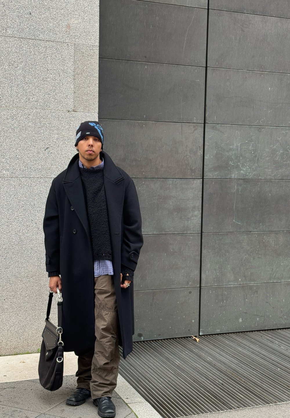 Άτομο που στέκεται μπροστά σε μια υφασμάτινη γκρι φόντο, φορώντας ένα μακρύ μαύρο παλτό, πλεκτό πουλόβερ, καρό πουκάμισο, φαρδιά καμηλό παντελόνια και ένα καπέλο με σχέδιο.