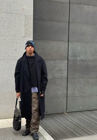 Άτομο που στέκεται μπροστά σε μια υφασμάτινη γκρι φόντο, φορώντας ένα μακρύ μαύρο παλτό, πλεκτό πουλόβερ, καρό πουκάμισο, φαρδιά καμηλό παντελόνια και ένα καπέλο με σχέδιο.