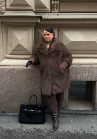 Brun fuskpälsjacka med bred krage, ribbstickade ärmar, tillsammans med bruna leggings och knähöga läderstövlar. Svart handväska på marken.