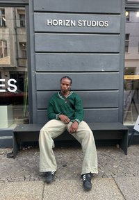 Πράσινο πουλόβερ με πλέξη καλωδίου και λευκές λεπτομέρειες, χαλαρές μπεζ παντελόνες και μαύρα αθλητικά παπούτσια. Καθισμένο σε ένα παγκάκι κοντά σε ένα κατάστημα.